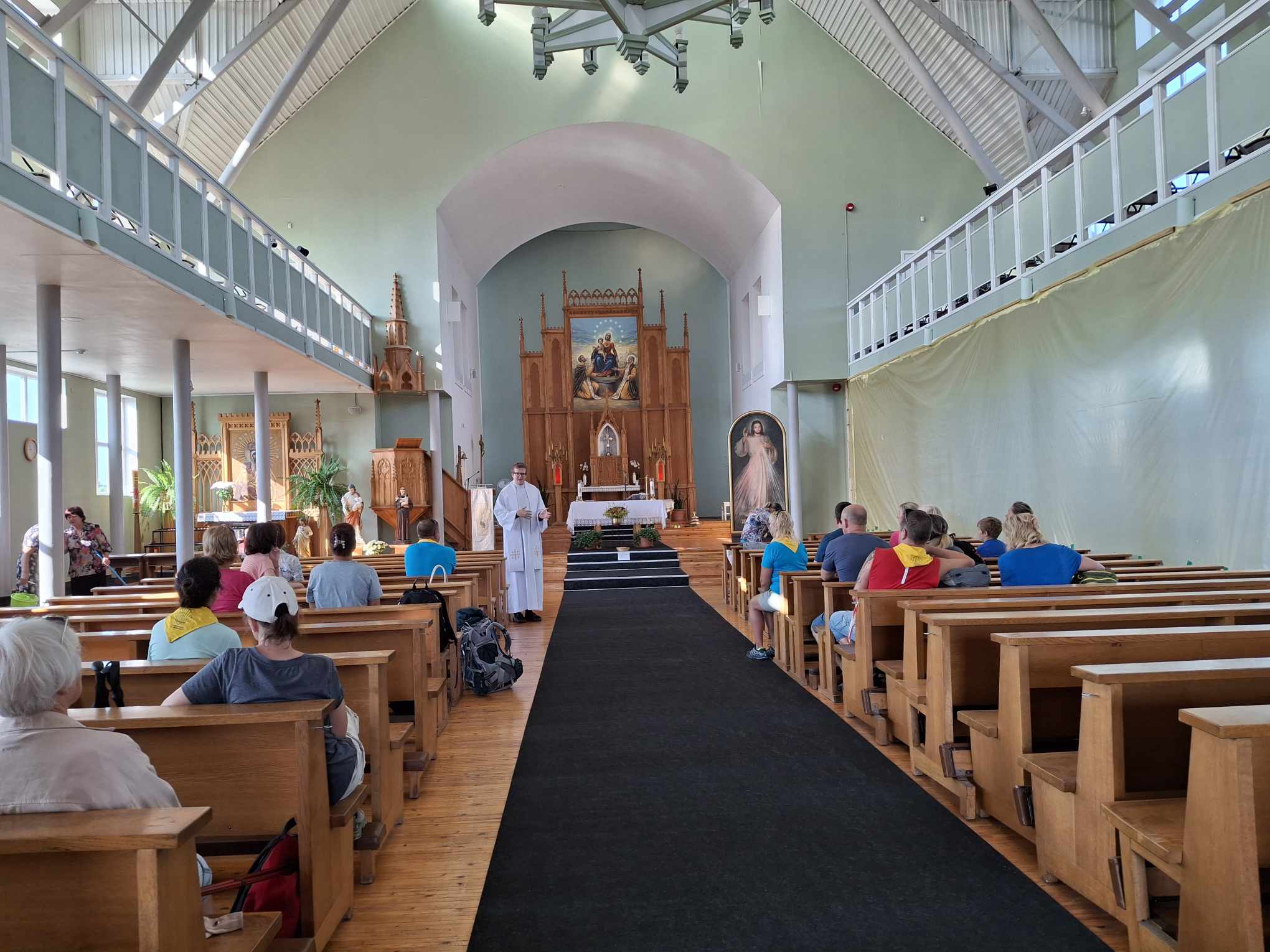 Baznīcas iekšpuse dievkalpojuma laikā. Priesteris baltās drēbēs runā ar draudzes locekļiem, kas sēž koka solos. Baznīcai ir augsti griesti un balkoni, bet labā siena daļēji nosegta ar baltu materiālu, iespējams, remonta dēļ.