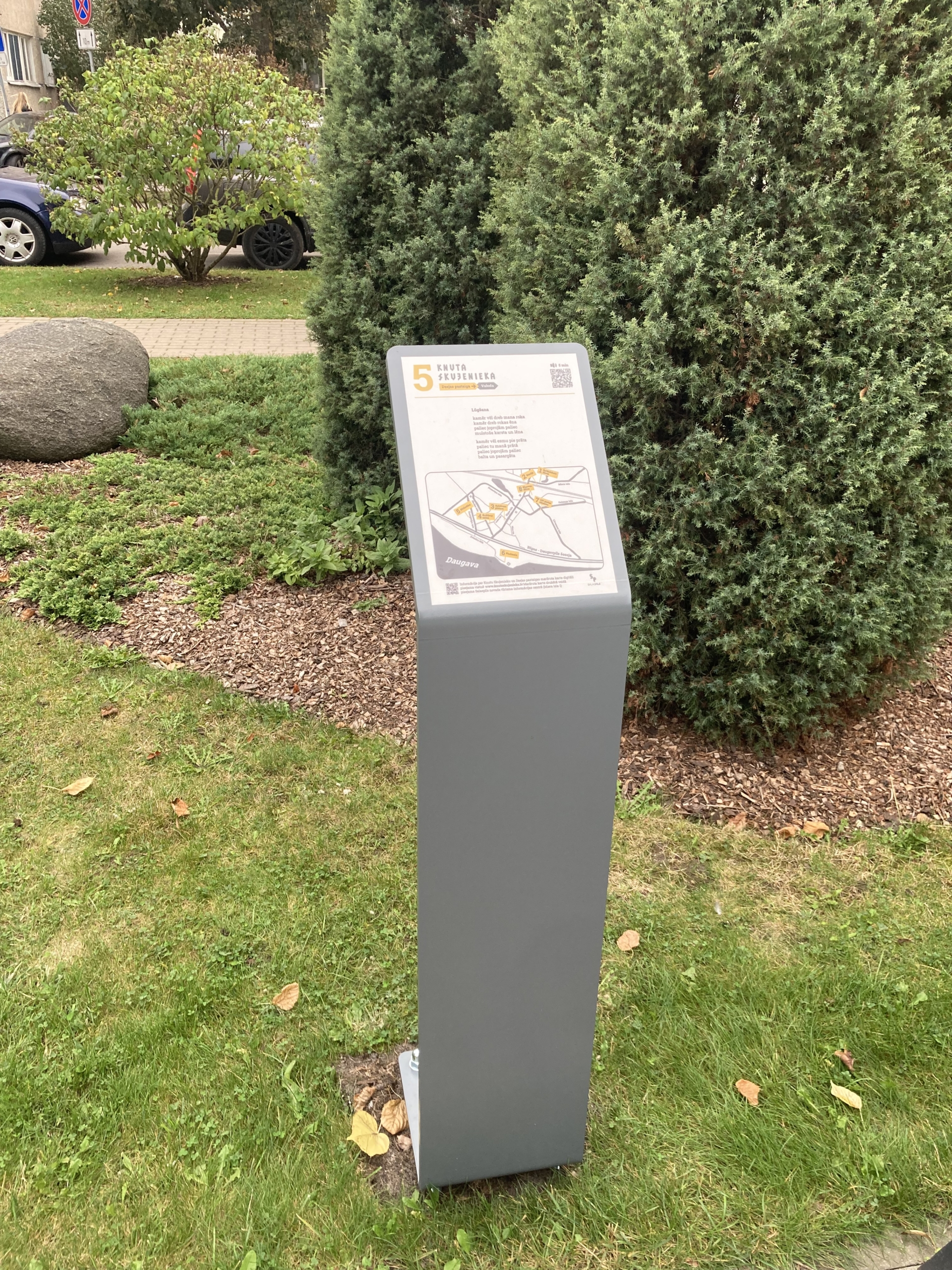 Pelēks informatīvs stends zālājā, uz kura redzama karte ar iezīmētām vietām, virsraksts "Knuta Skujenieka dzejas pastaiga" un QR kods.