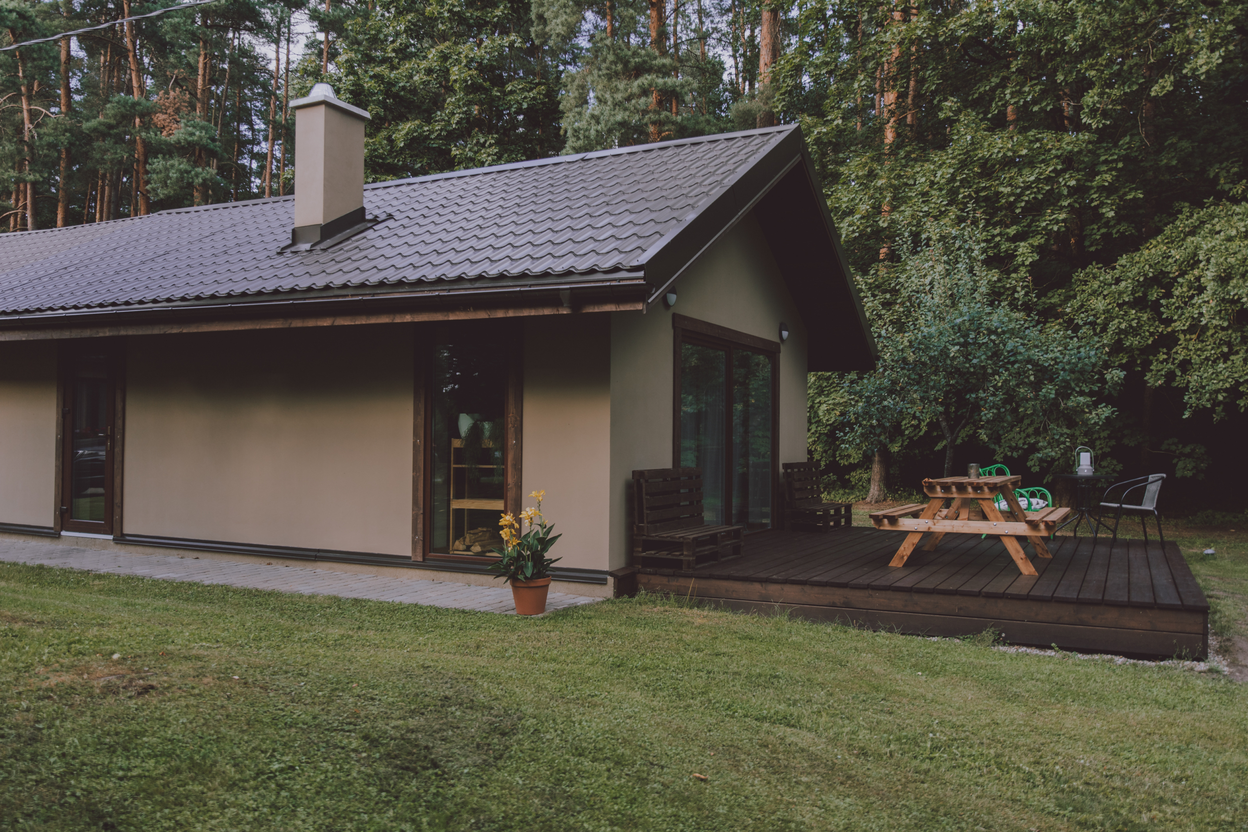 Moderna, gaišas krāsas māja ar brūnu jumtu un skursteni, pie kuras atrodas koka terase ar piknika galdu un soliem. Apkārtne ir klāta ar zaļu zāli, bet fonā redzams blīvs mežs.