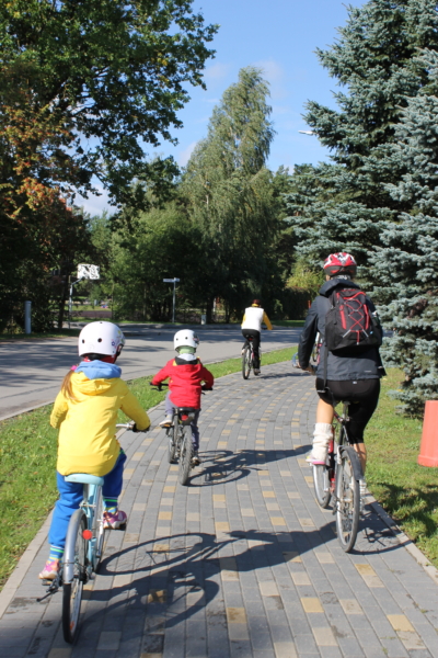 Ģimene Salaspilī brauc ar velosipēdiem pa bruģētu veloceliņu, ko ieskauj koki, izbaudot aktīvu atpūtu dabā.
