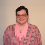 Liga-Dziedataja Gide Līga Dziedātāja - smaidīga sieviete ar brillēm, rozā kreklā un rūtoti rozā jakā.