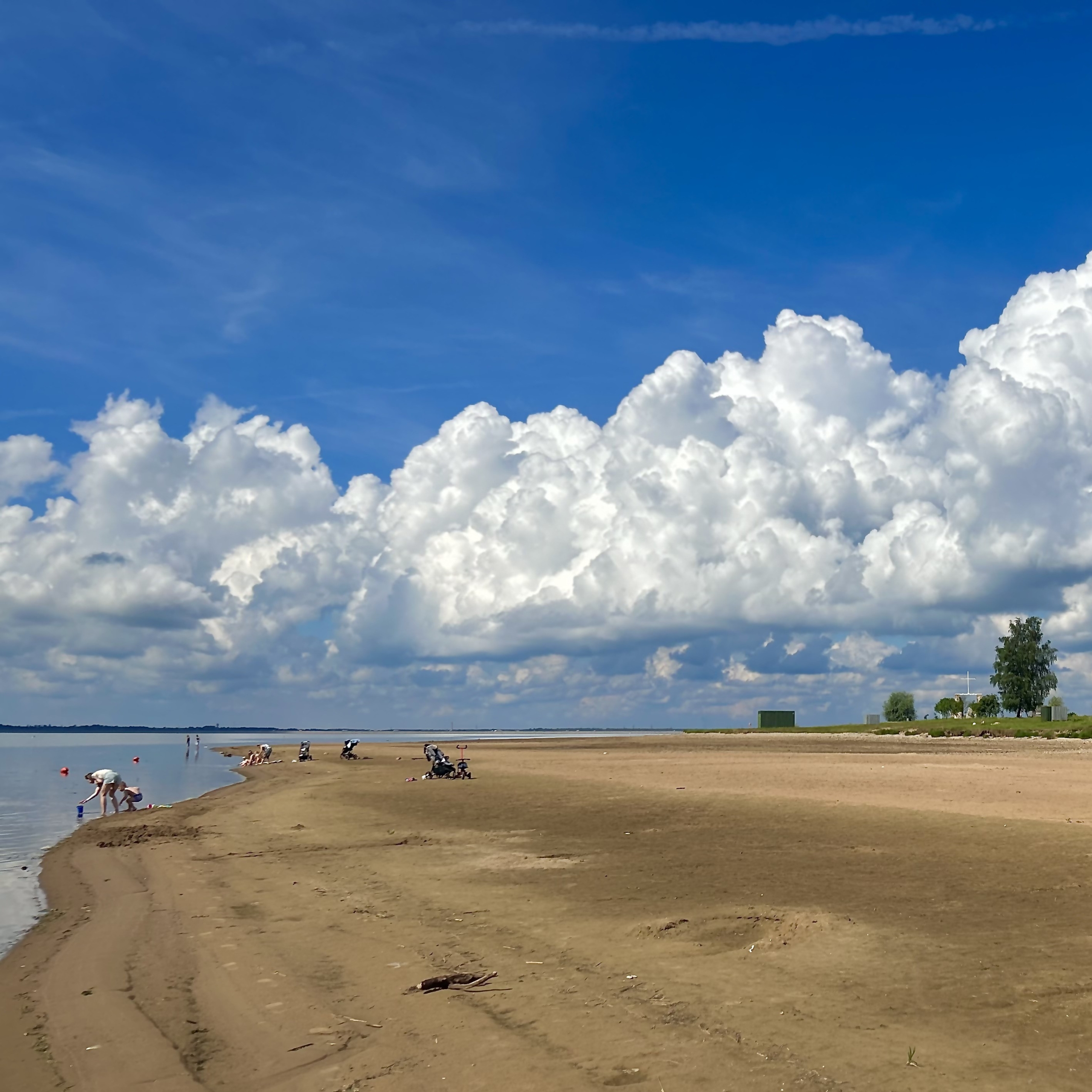 Saulaina diena Salaspils pludmalē ar zilu debesi un lieliem baltiem mākoņiem. Cilvēki atpūšas pie ūdens un gar krastu.