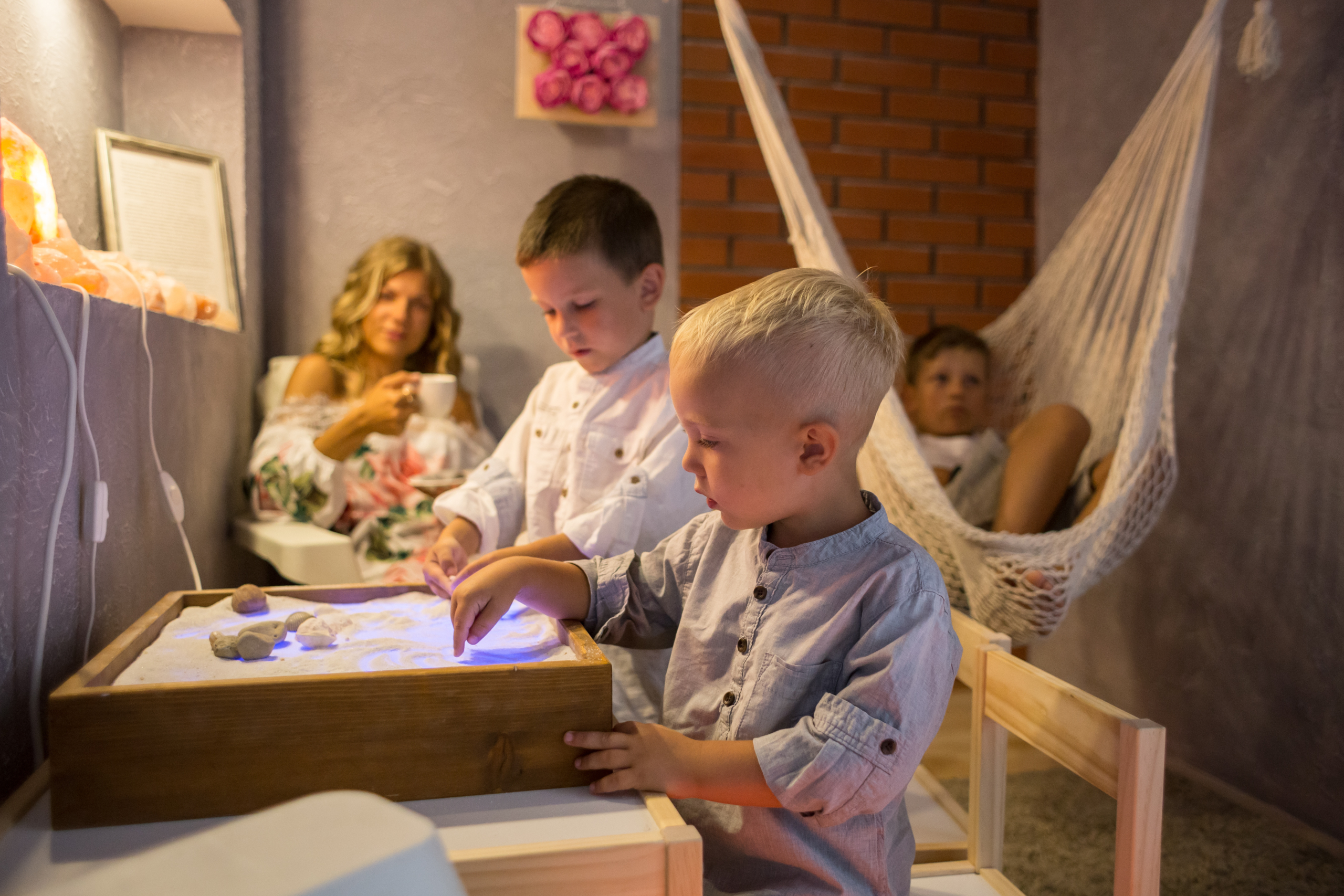 Ģimene atpūšas Salaspils sāls istabā, kur bērni rotaļājas ar apgaismotu smilšu galdu, kamēr pieaugušie bauda mierīgu atmosfēru ar sāls lampām un šūpuļtīklu.