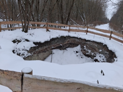 Sniegots liels zemes iegruvums - Salaspils karsta kritene ar redzamiem zemes slāņiem, norobežots ar koka sētu ziemīgā mežā.