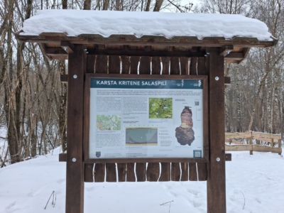 Koka informācijas stends, uz kura lasāms uzraksts "KARSTA KRITENE SALASPILĪ", atrodas sniegotā ziemas ainavā.