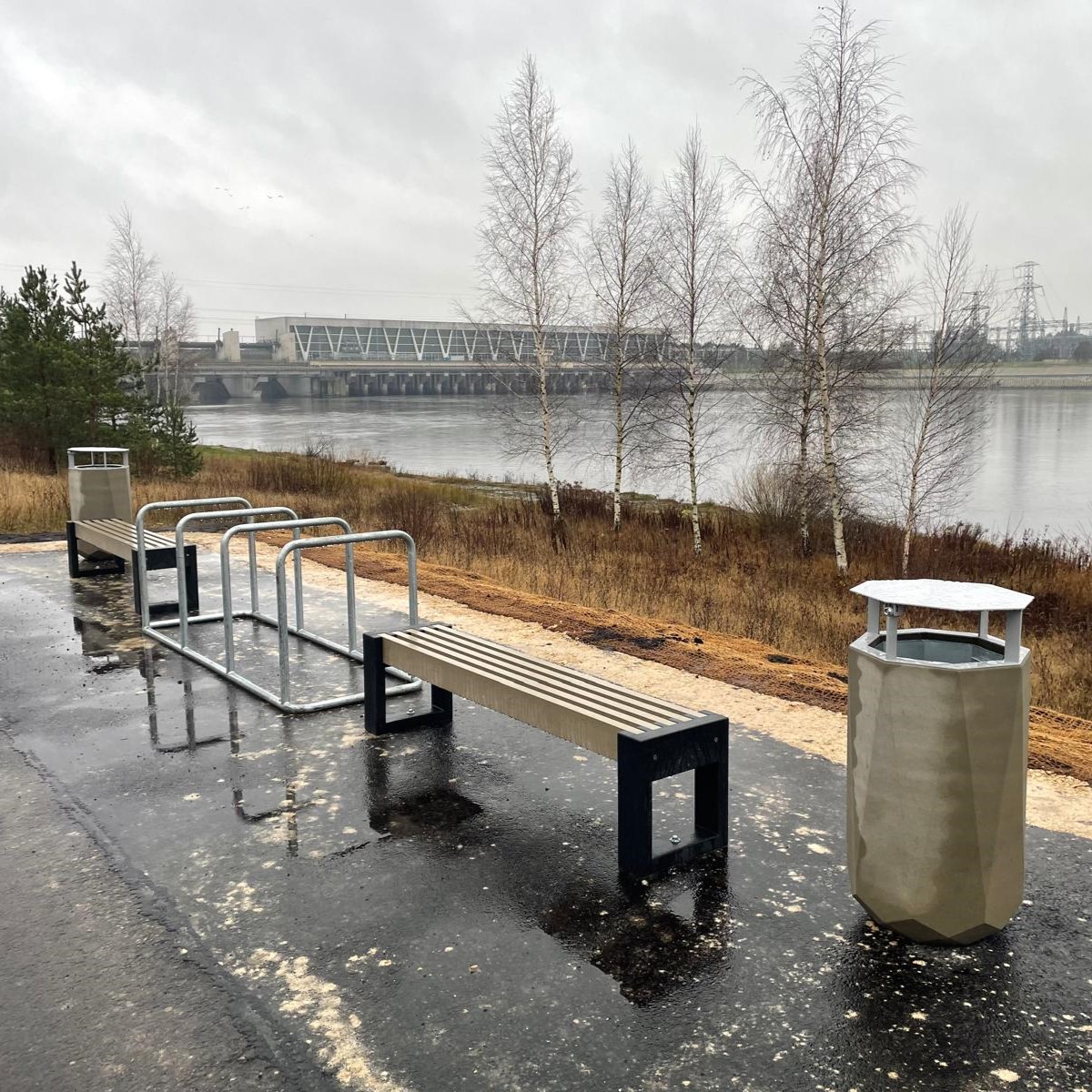 Asfaltēts celiņš ar soliņiem un velosipēdu statīvu Zirņu salā, ar skatu uz Rīgas hidroelektrostaciju.