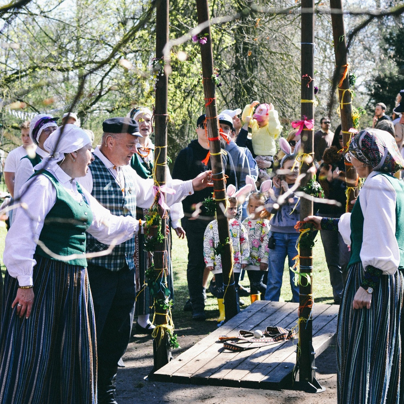 Pavasarīgs Lieldienu pasākums Daugavas muzejā, kur cilvēki tautastērpos pulcējas ap izrotātām Lieldienu šūpolēm, radot svinīgu un tradicionālu atmosfēru.