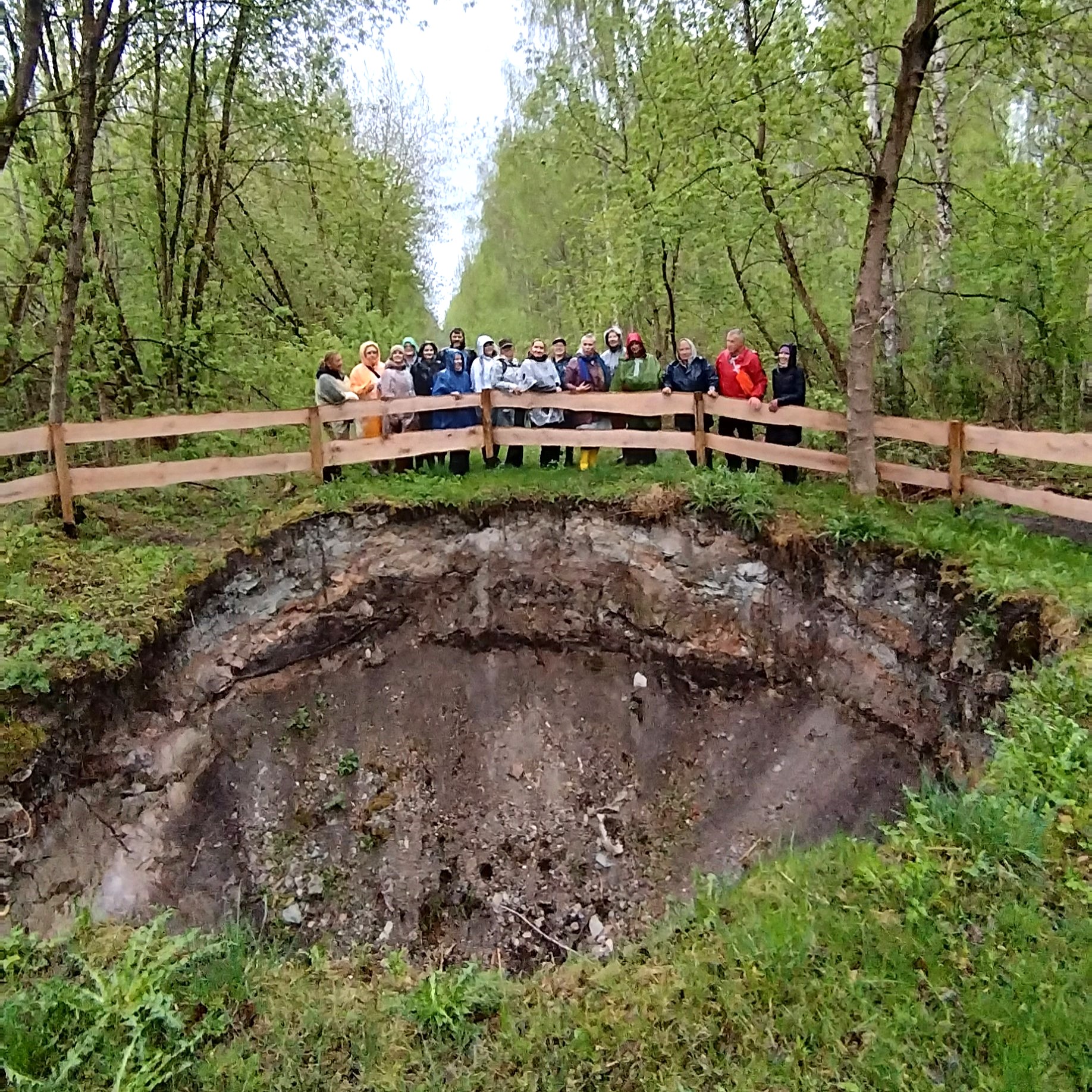 Cilvēku grupa stāv pie koka žoga, vērojot Salaspils karsta kriteni meža ieskautā vietā.